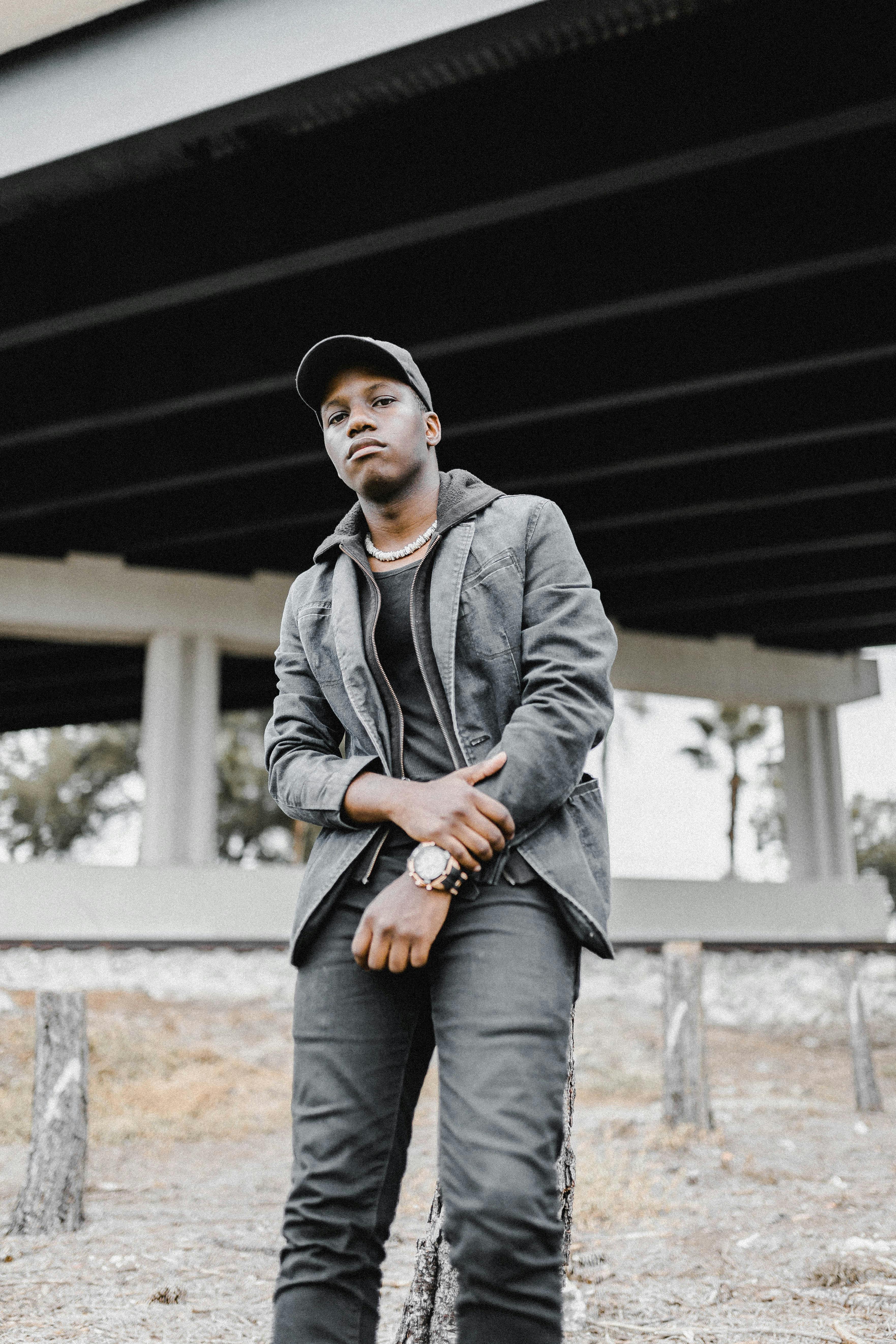 Stylish young man in a black outfit posing confidently under an urban bridge.