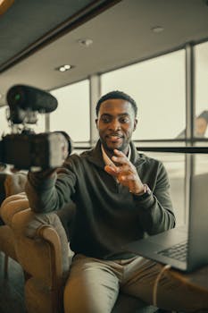 A man broadcasting with a camera and laptop indoors, creating live content in a modern space.
