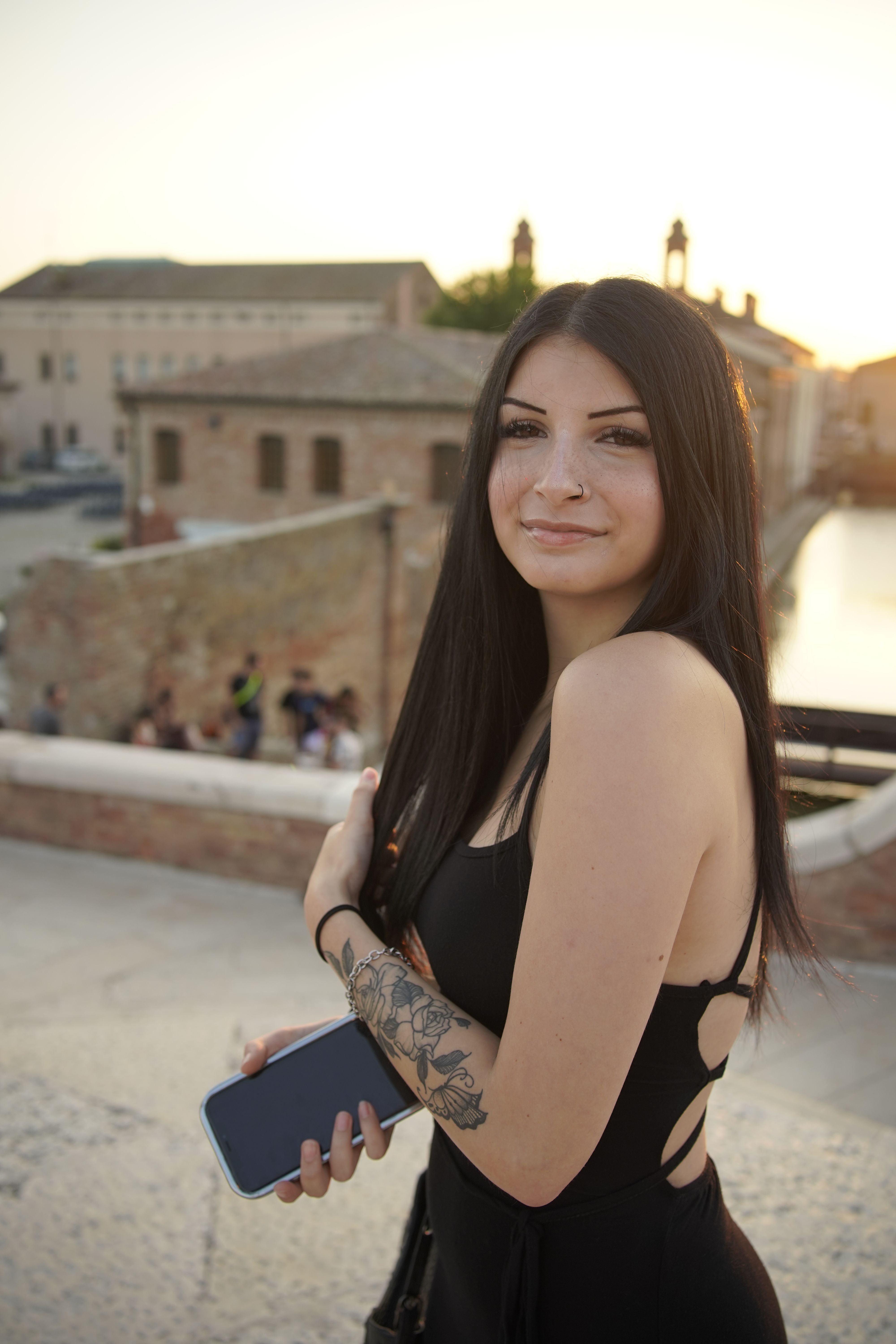 Young woman in black dress holding smartphone by Comacchio canal at sunset.