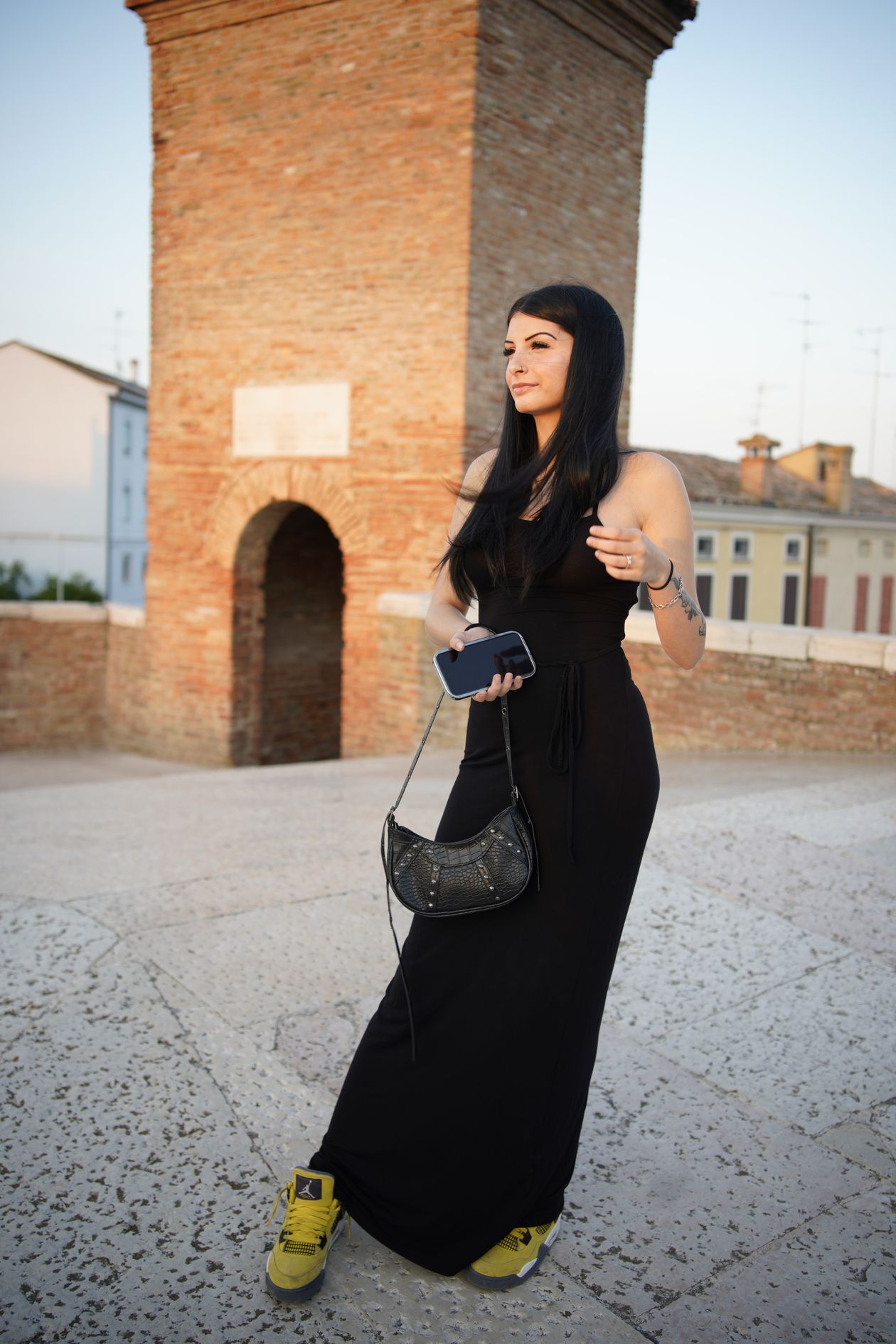 Woman in a long black dress and yellow sneakers standing by a brick tower.