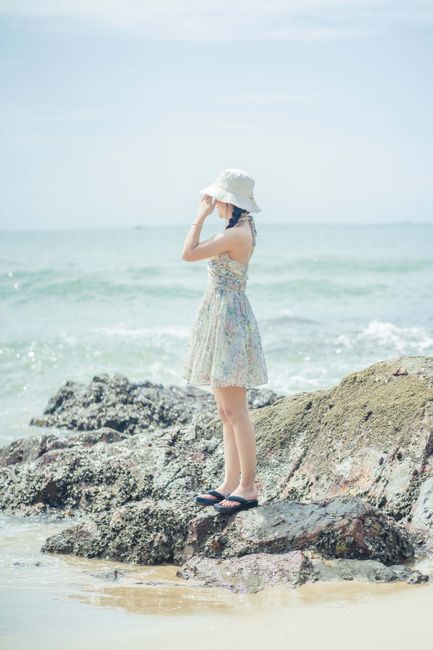 A woman in a floral dress and bucket hat stands on rocks by the sea, enjoying a sunny day.