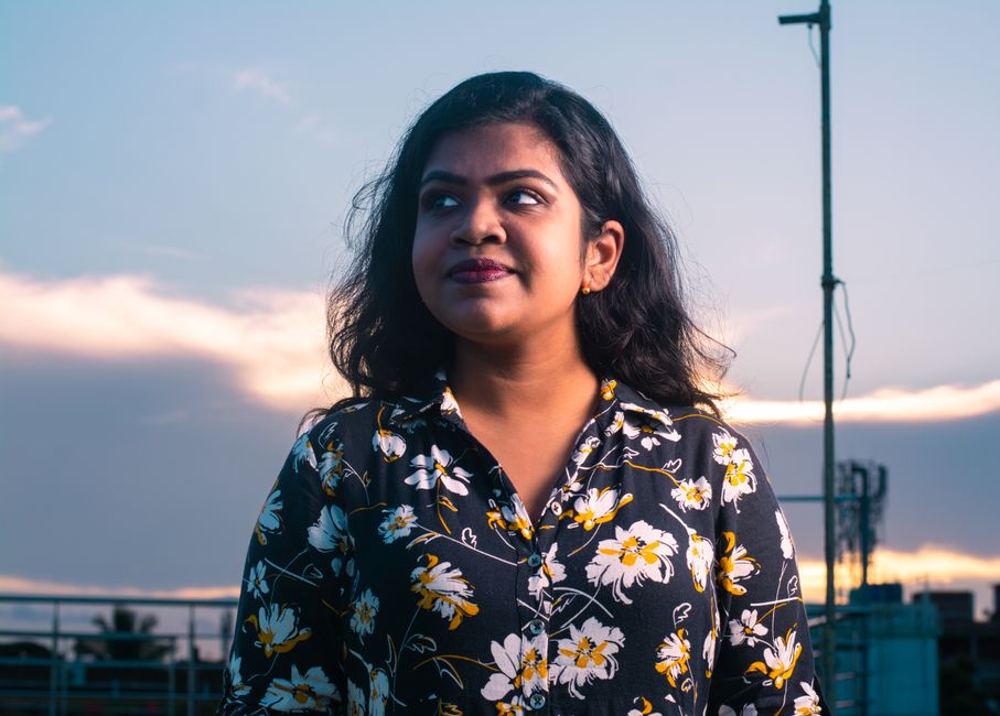 A thoughtful woman in a floral shirt stands outdoors at sunset, gazing upward.