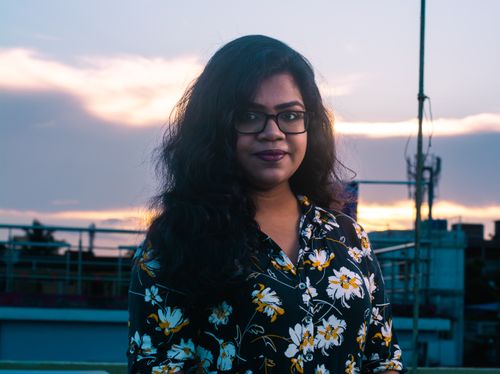 Free A woman in glasses and floral shirt poses on a rooftop at dusk, with an urban skyline behind her. Stock Photo