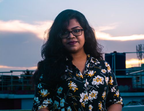 Free A young woman with eyeglasses and a floral shirt smiles on a terrace at sunset. Stock Photo