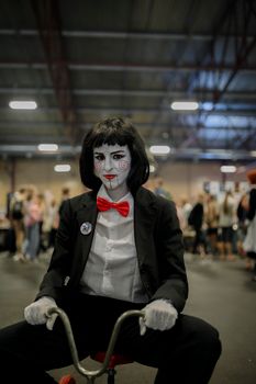 A person in a Saw-inspired cosplay costume sitting inside a bustling indoor event.