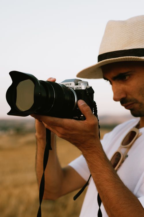 Free A man in a hat intently photographs a scene outdoors with a professional camera. Stock Photo