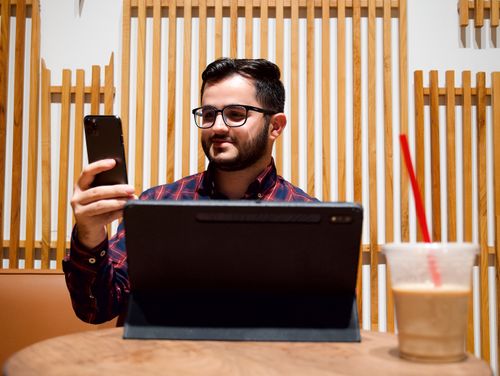Free Adult man with glasses using smartphone and tablet at a cafe table with a drink. Stock Photo