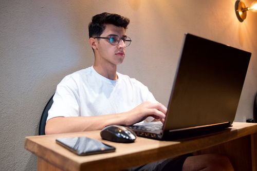 Free Man in white shirt using laptop at desk indoors with smartphone and mouse nearby. Stock Photo