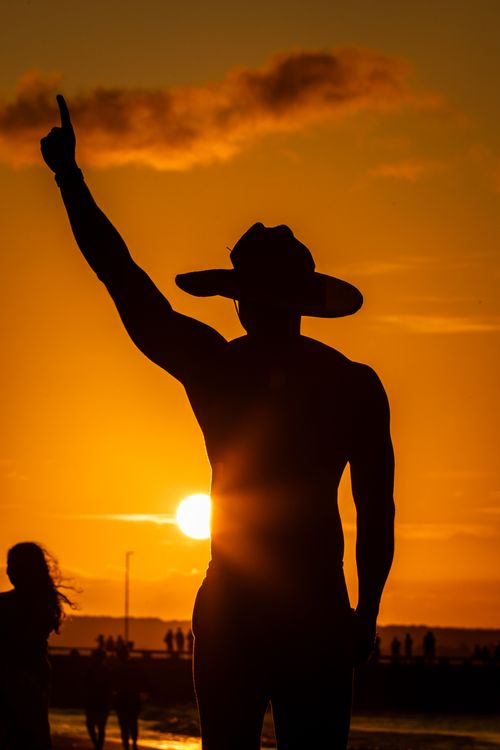 Free Silhouette of a man raising his arm against a vibrant sunset sky, wearing a hat, with people in the background. Stock Photo