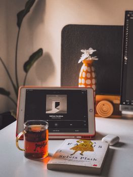 A cozy setup with a tablet playing music, tea, and a book on a desk by the window.