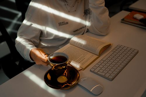 Free A woman reading a book while holding a cup of tea at a desk with sunlight streaming in. Stock Photo