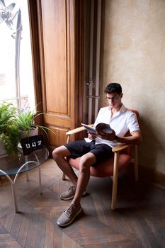 A young man in a white shirt reads a book in a cozy room with plants and natural light.