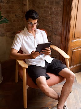 A young man sits in an armchair reading a tablet inside a sunlit room.