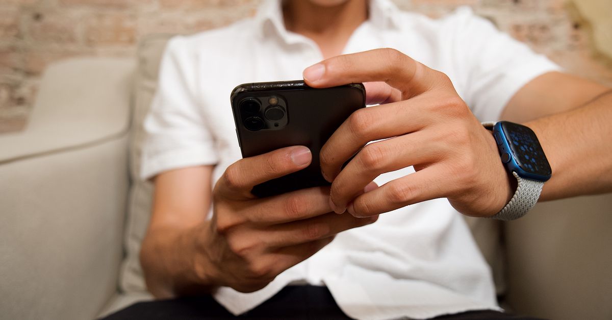 A person sitting on a couch staring at a phone while a clock on the wall shows 8pm, dim lighting