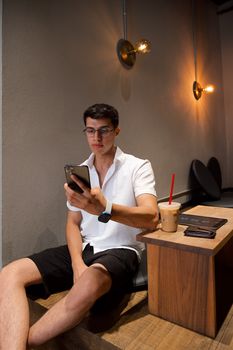 A young man enjoying his time in a stylish café, using a smartphone with a drink on the table.