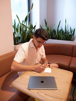A man with glasses writes in a notebook indoors next to a laptop in a coffee shop.