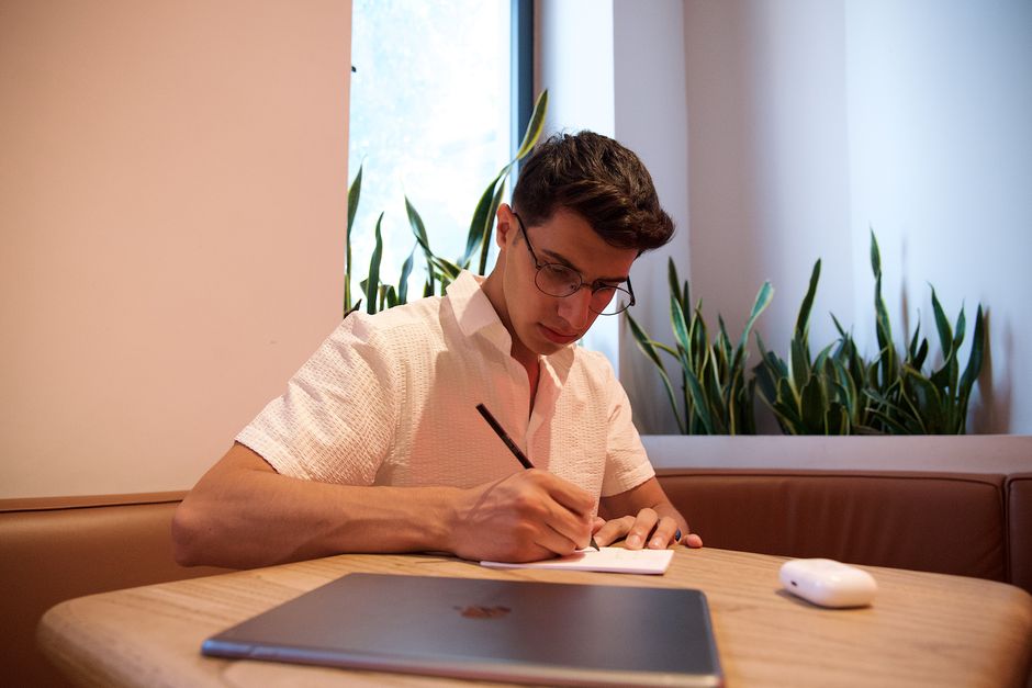 A young adult writing notes in a cafe with a laptop and plants in the background. A young adult writing notes in a cafe with a laptop and plants in the background.