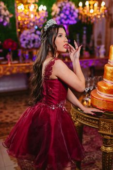 Beautiful quinceañera girl posing in a lavish burgundy dress with elegant decor.