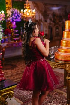 Teenage girl at her quinceañera holding a red rose in a luxurious setting with a golden cake.