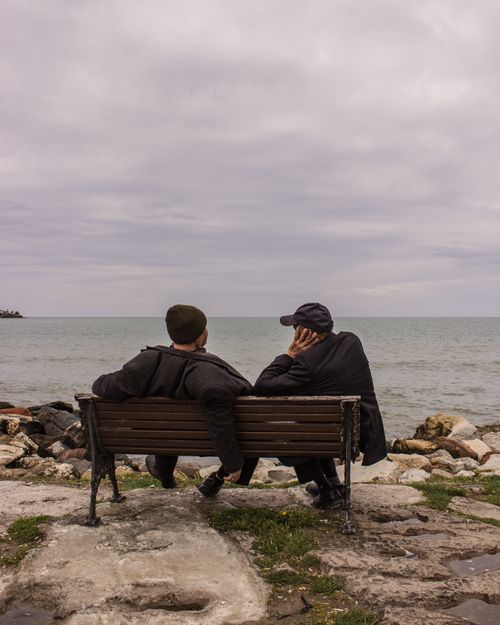 Free Two men sit and talk on a bench overlooking the rocky Black Sea coast in Ünye, Türkiye. Stock Photo