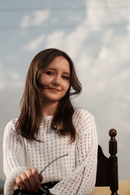 Free Stylish young woman in white sweater poses outdoors, exuding elegance and charm against a cloudy sky. Stock Photo
