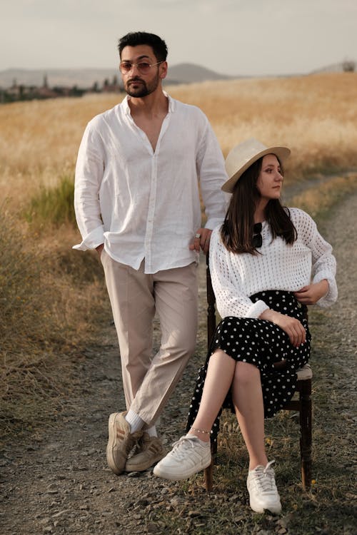 Woman in a White Sweater and Black Polka Dot Skirt Sitting Next to a Man in a White Shirt and Beige Pants Standing on a Dirt Road