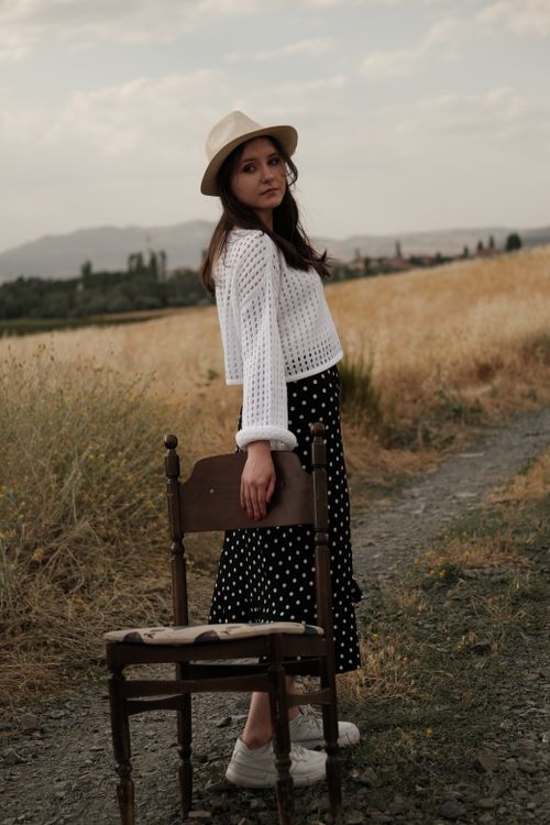 Free Fashionable woman in hat and polka dot skirt standing by chair in sunny meadow. Stock Photo