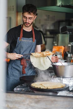 A focused male chef wearing an apron makes crepes in a kitchen setting, Saint-Nazaire, France.