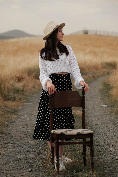 Model in a White Sweater and Fedora Standing with a Chair on a Dirt Road
