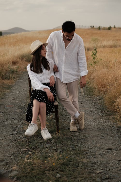 Woman in a White Sweater and Midi Skirt Sitting Next to a Man in a White Shirt and Beige Pants Standing on a Dirt Road