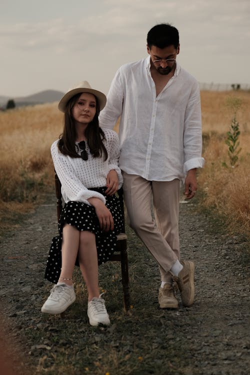 Man in a White Shirt and Beige Pants Standing Next to a Woman in a White Sweater and Black Skirt Sitting on a Chair Among the Fields