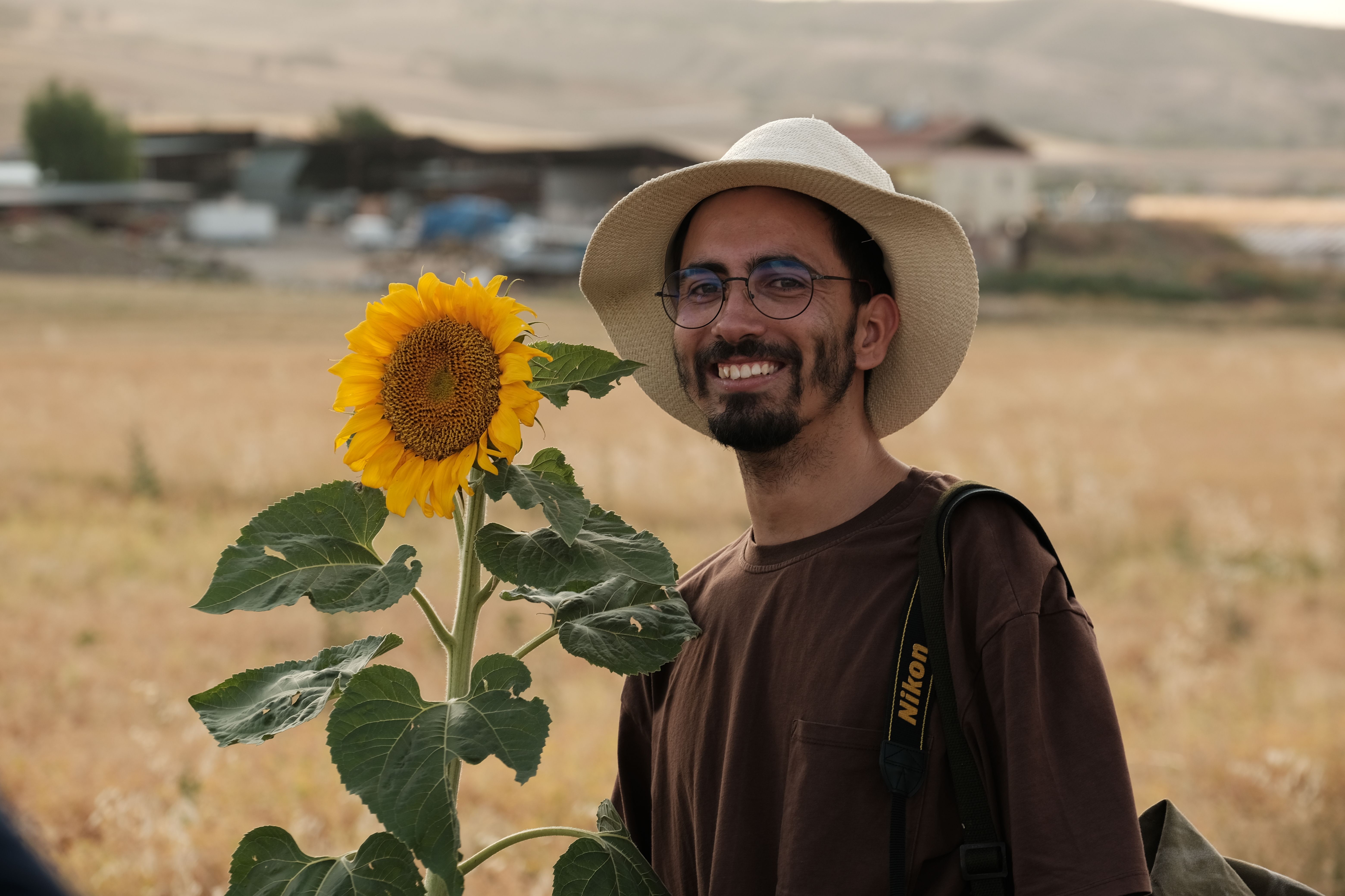 A cheerful man wearing a sunhat and glasses poses with a sunflower in a rural landscape.