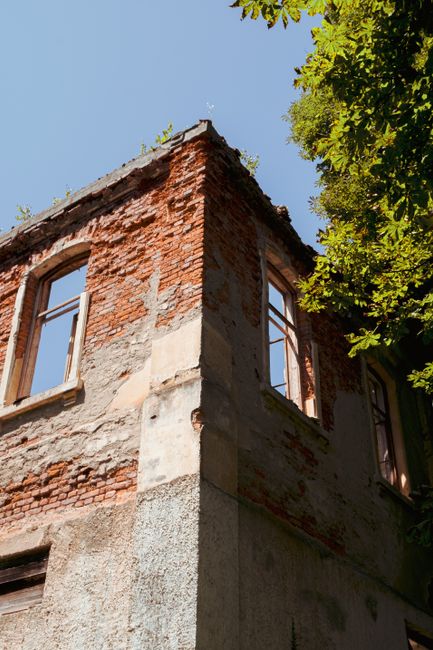 Ruined brick building facade with open windows under clear sky and tree shade.