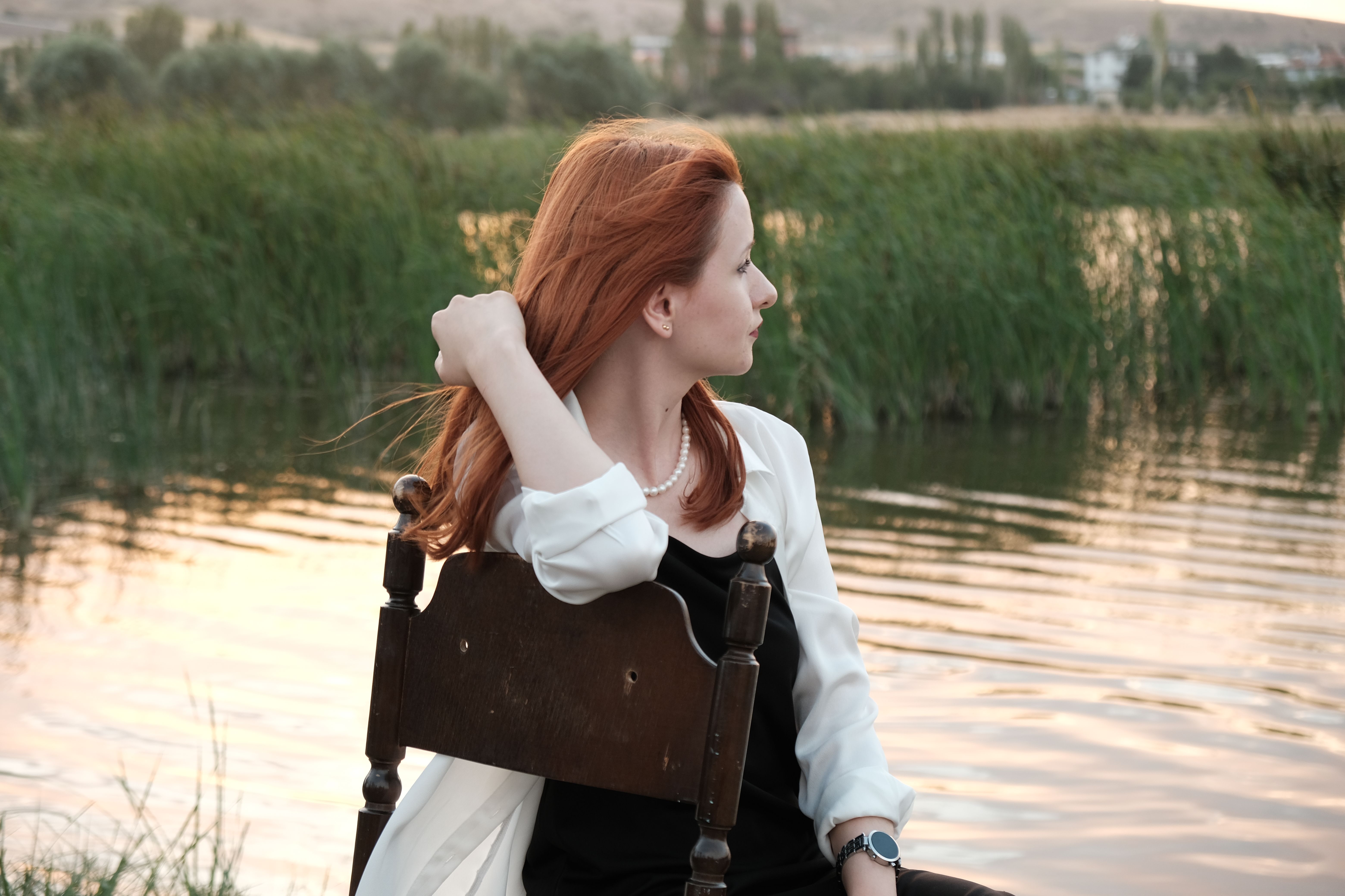 A red-haired woman in fashion attire sits by a serene pond, reflecting during sunset.