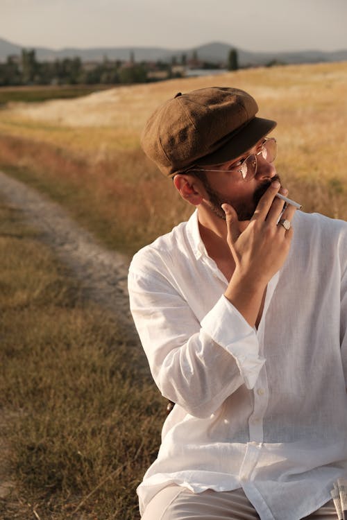 Model in a Newsboy Cap and White Shirt Smoking a Cigarette on a Dirt Road Through the Fields