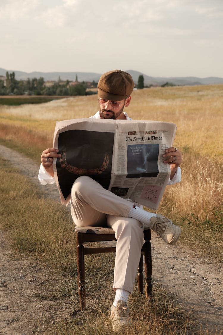 Elegant Man Sitting On A Chair On A Rural Field And Reading A Newspaper 