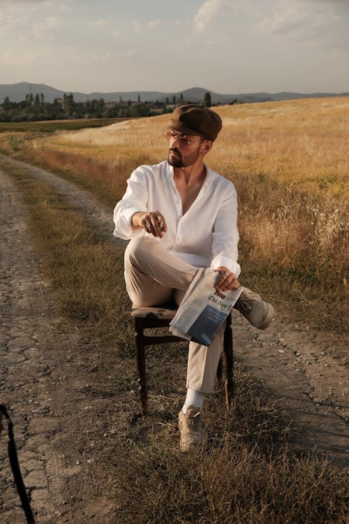 Man in a Newsboy Cap of a White Shirt and Beige Pants Sitting on a Chair in the Middle of a Dirt Road