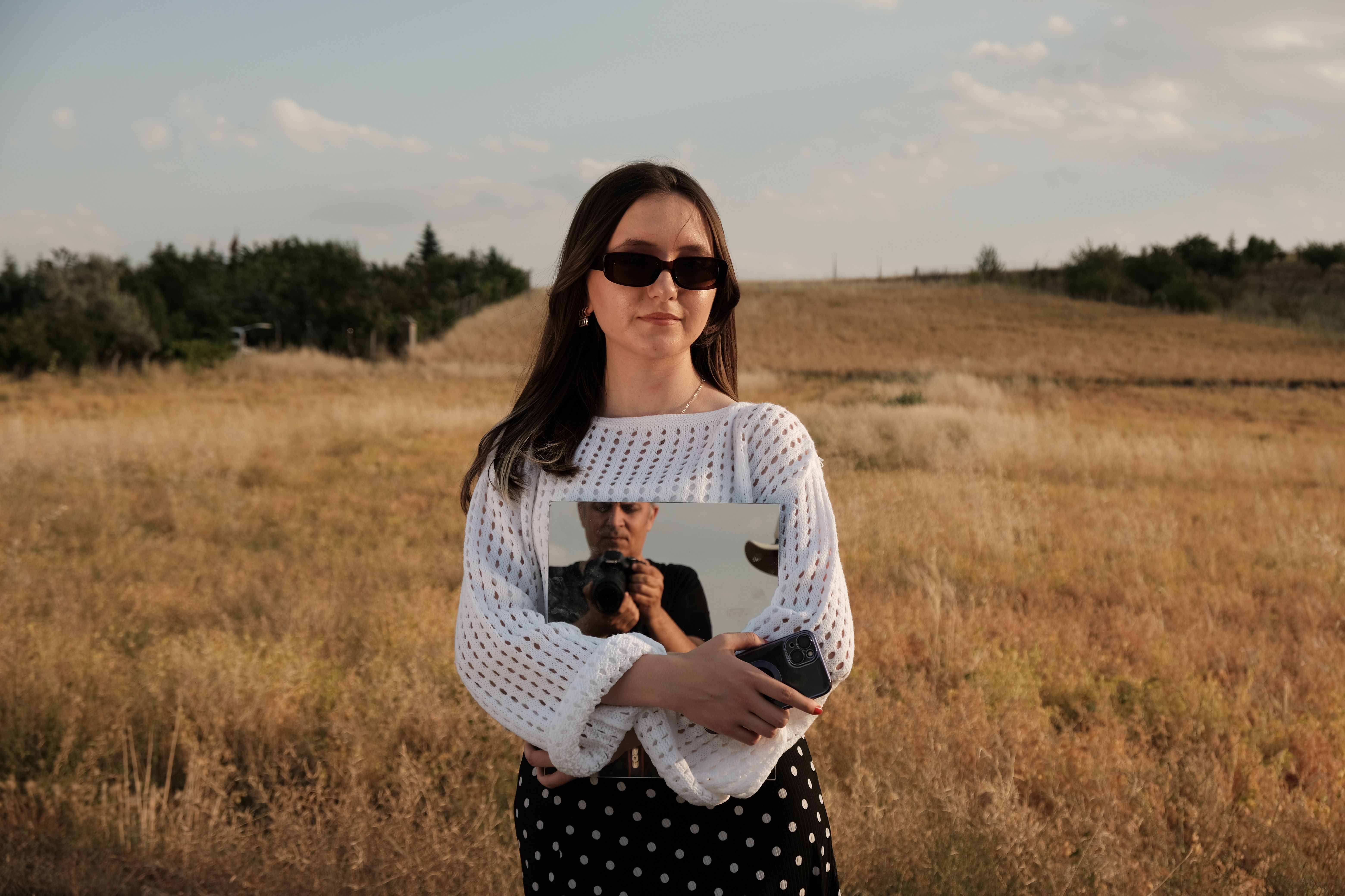 A woman stands in a field holding a mirror, reflecting a landscape view under a clear sky.