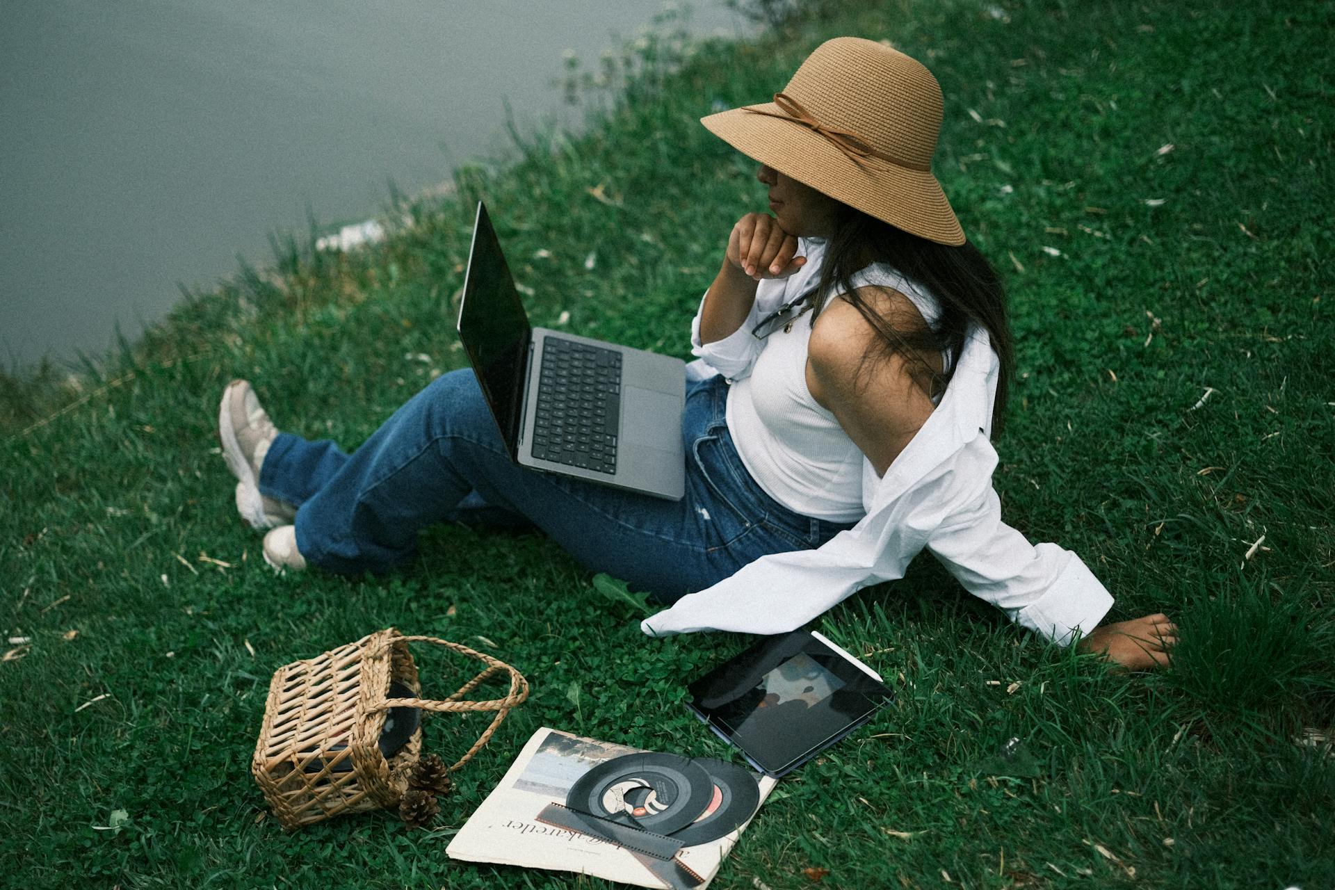 A woman sitting on grass by a lake working on a laptop, surrounded by leisure items.