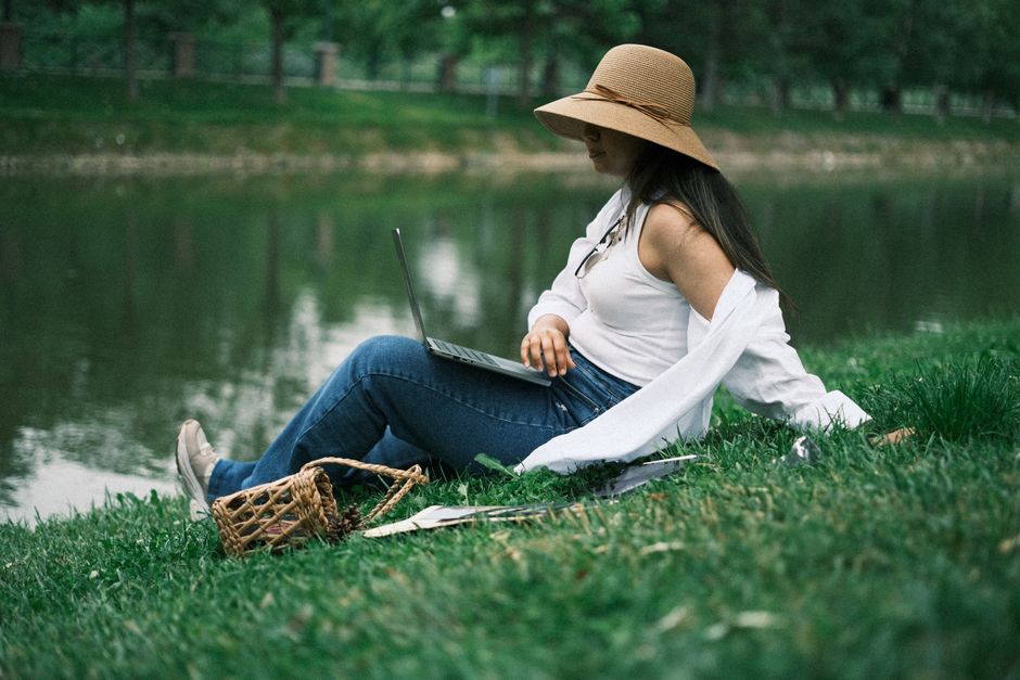 A woman using a laptop on a grassy park by a lake, working remotely in summer.