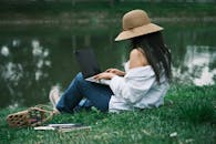 A Woman Sitting on the Ground by the River and Using a Laptop