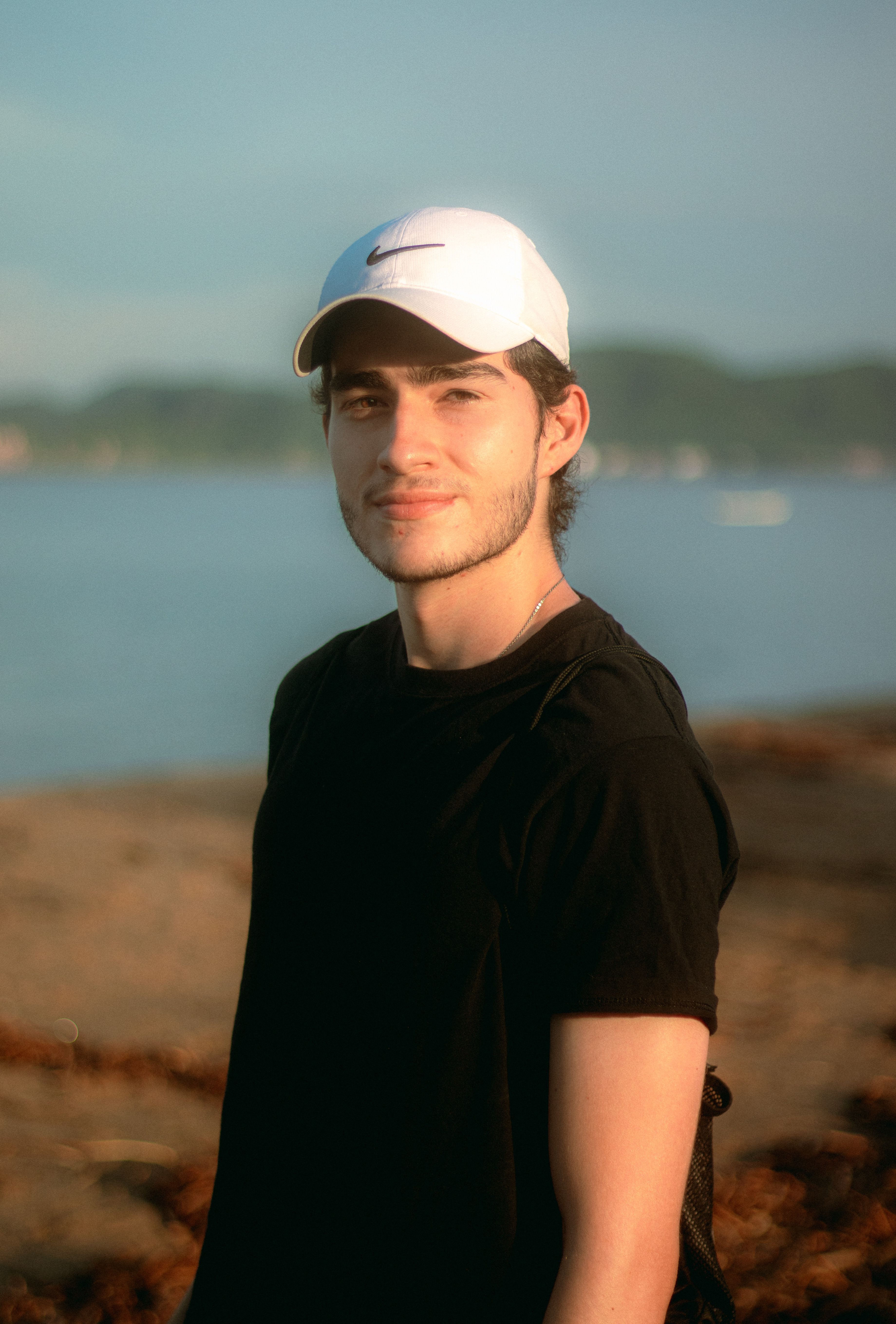 Portrait of a young man in a white cap and black t-shirt standing on a beach.