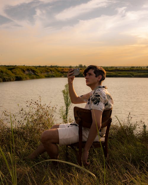 Young Man Sitting on the Riverbank Filming the Sunset