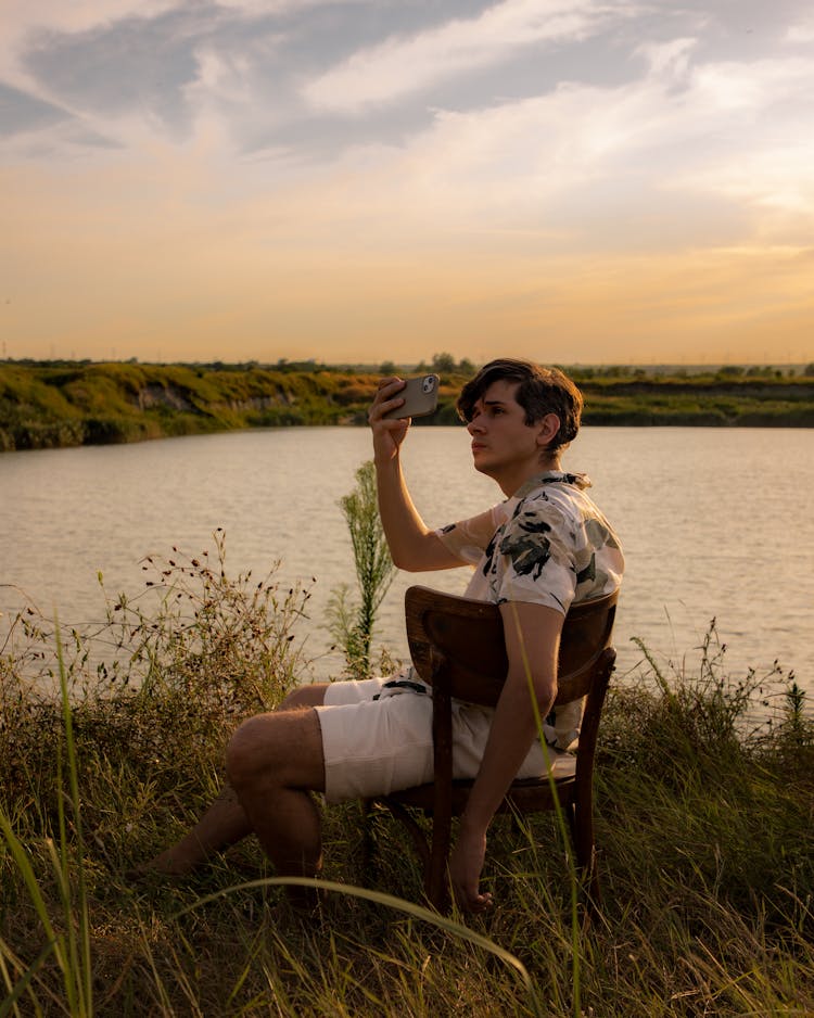 Jovan Vasiljević (a Young Man) Sitting On A Chair In The Middle Of A Meadow Holding A Phone, Taking A Photo Of The Sunset