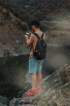 A man with a backpack photographs a mountain valley using a smartphone, showcasing an adventurous hiking experience.