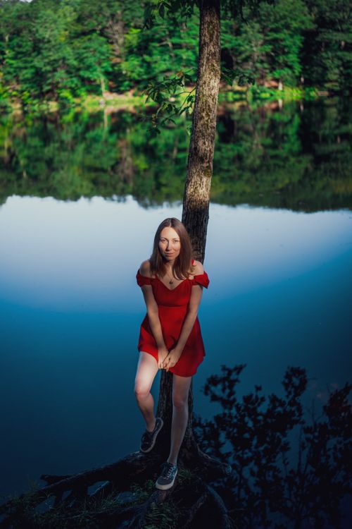 Free A woman in a red dress standing by a serene river in a lush green forest. Stock Photo