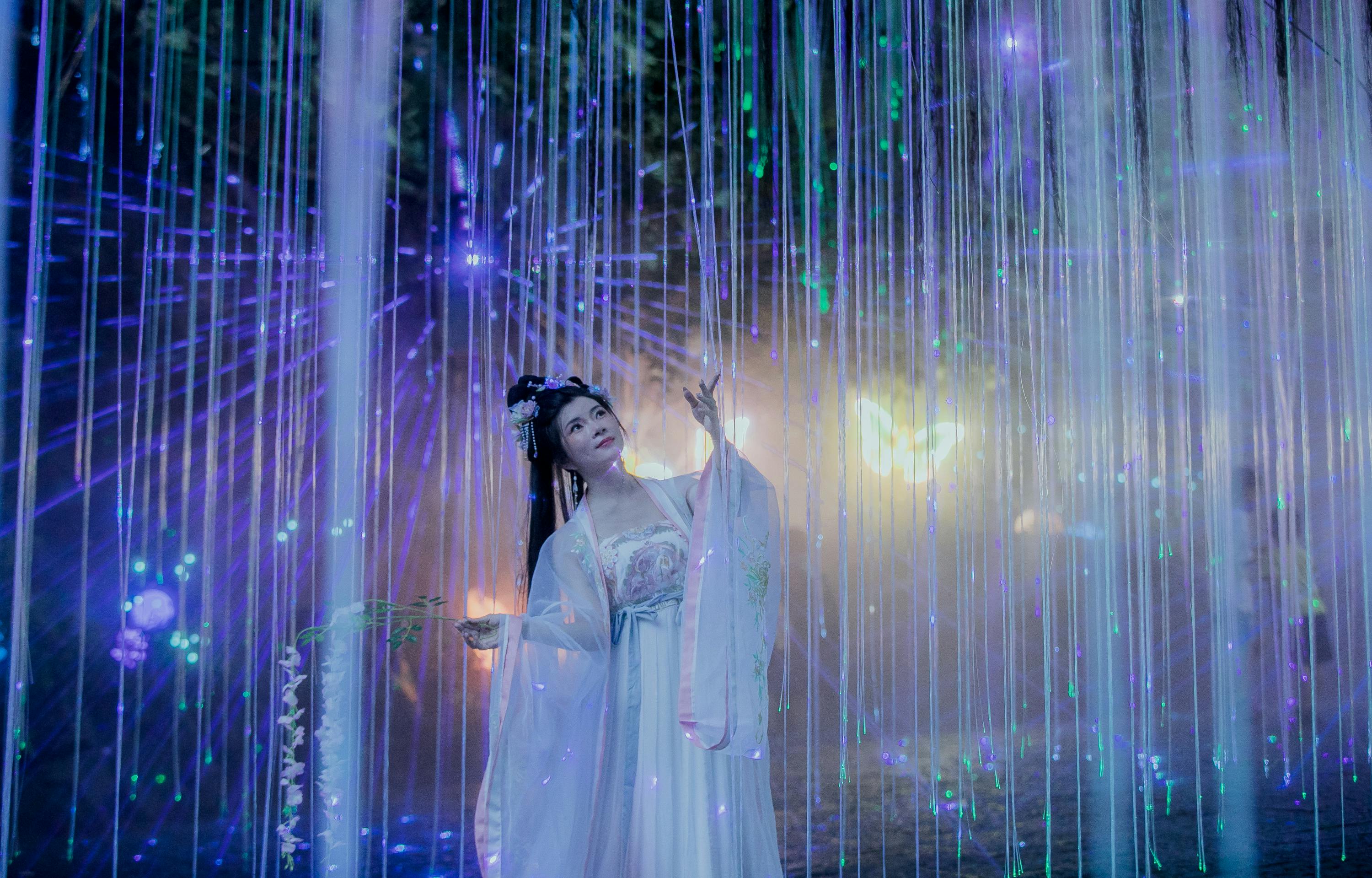 An Asian woman dressed in traditional attire, immersed in an illuminated fantasy scene with vibrant lights.
