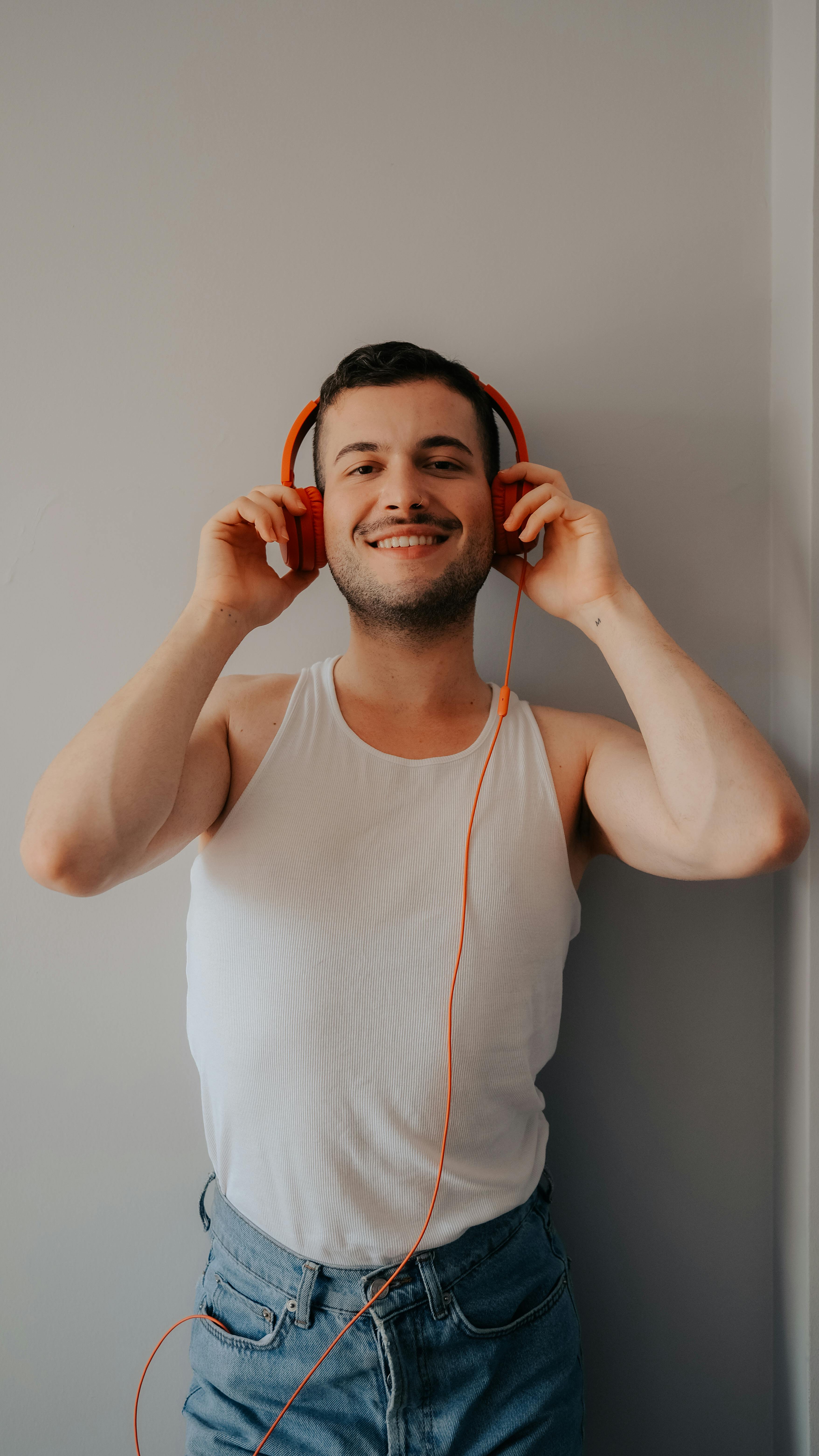 Happy man in a white tank top wearing orange headphones, enjoying music indoors.