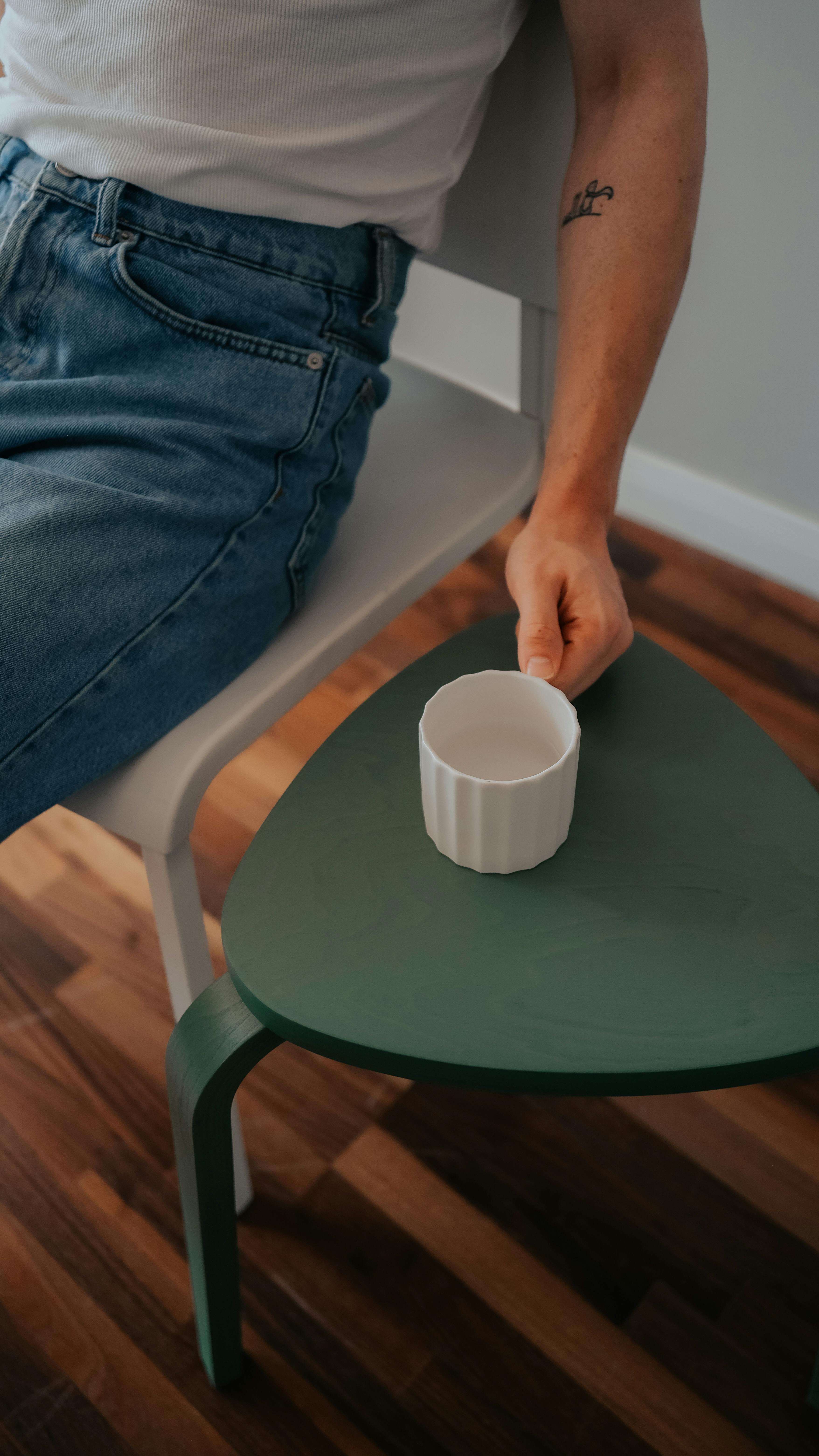 A person sits holding a mug on a green table with a wooden floor indoors.