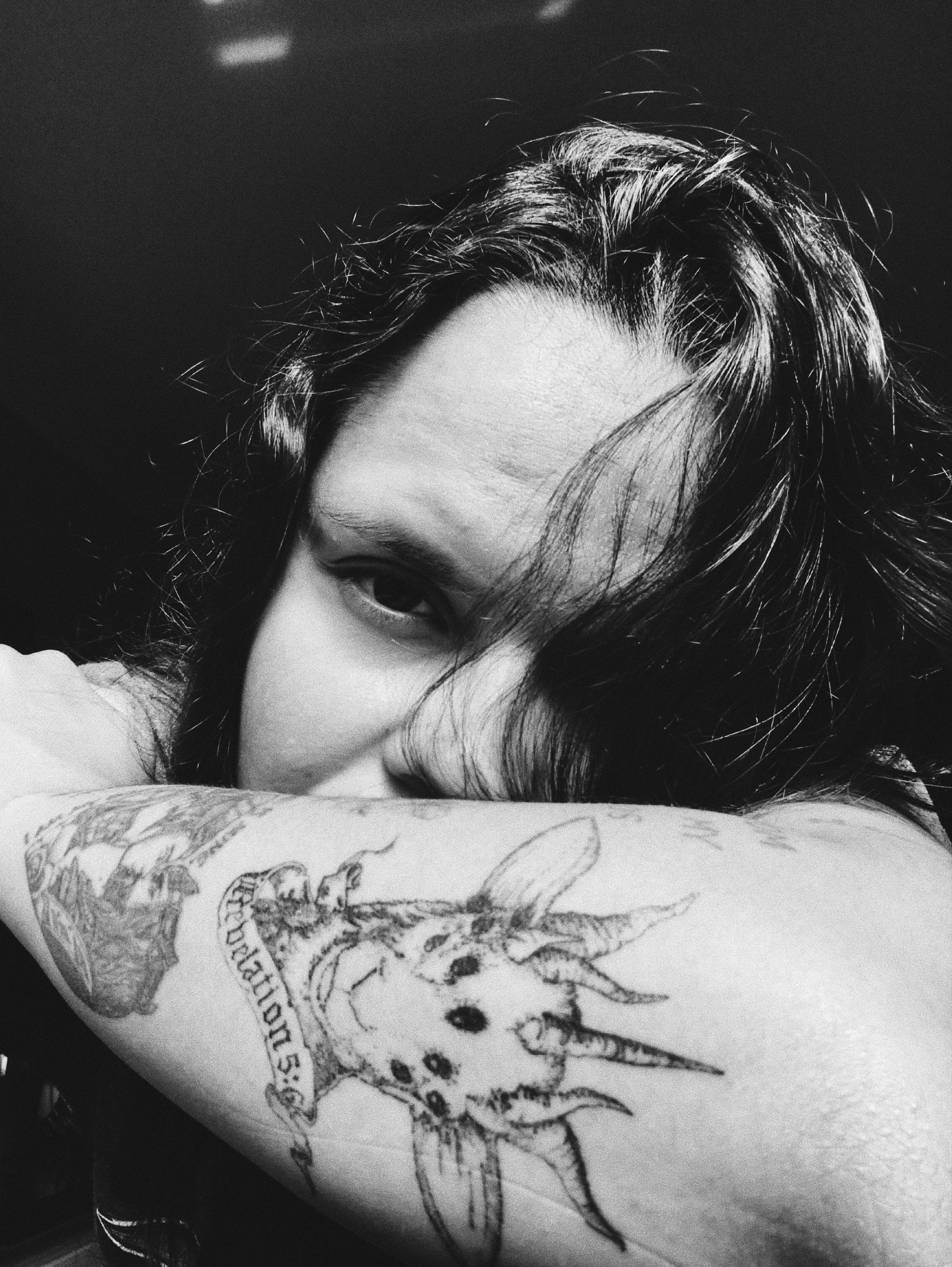 A thoughtful black and white portrait of a person with visible tattoos and long hair, showcasing introspection.